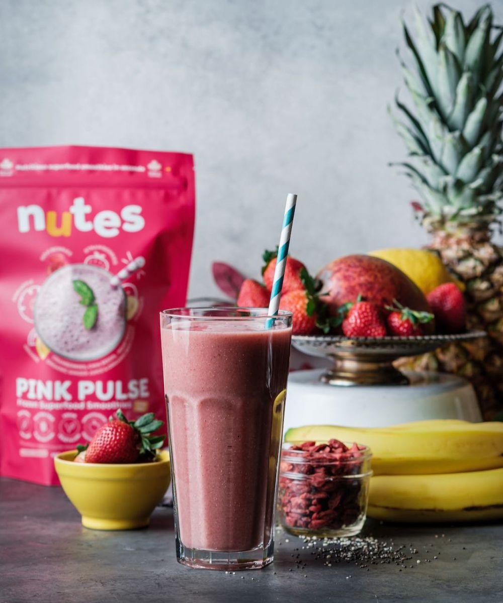 Nutes frozen superfood pink pulse Smoothie in glass with paper straw, surrounded by fresh fruit, berries, and product pouch.