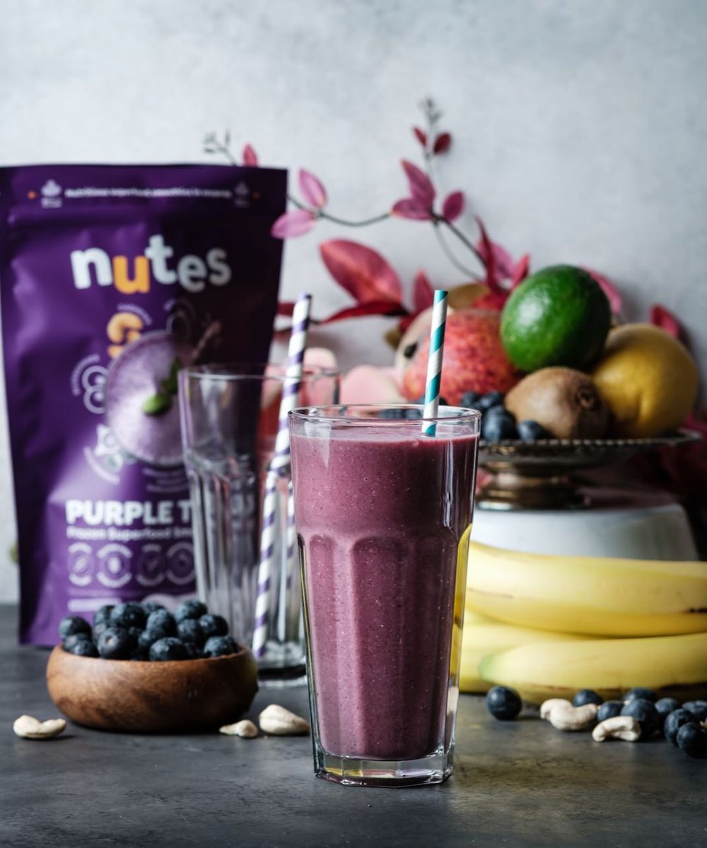 Nutes frozen superfood purple tonic Smoothie in glass with paper straw, surrounded by fresh fruit, berries, and product pouch.