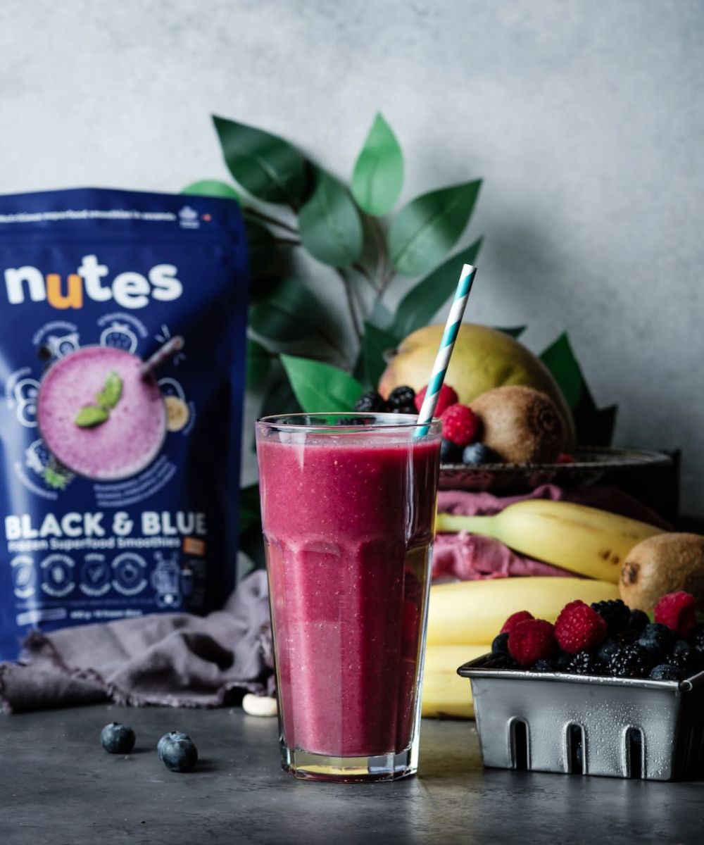 Nutes frozen superfood black & blue Smoothie in glass with paper straw, surrounded by fresh fruit, beans, and product pouch.