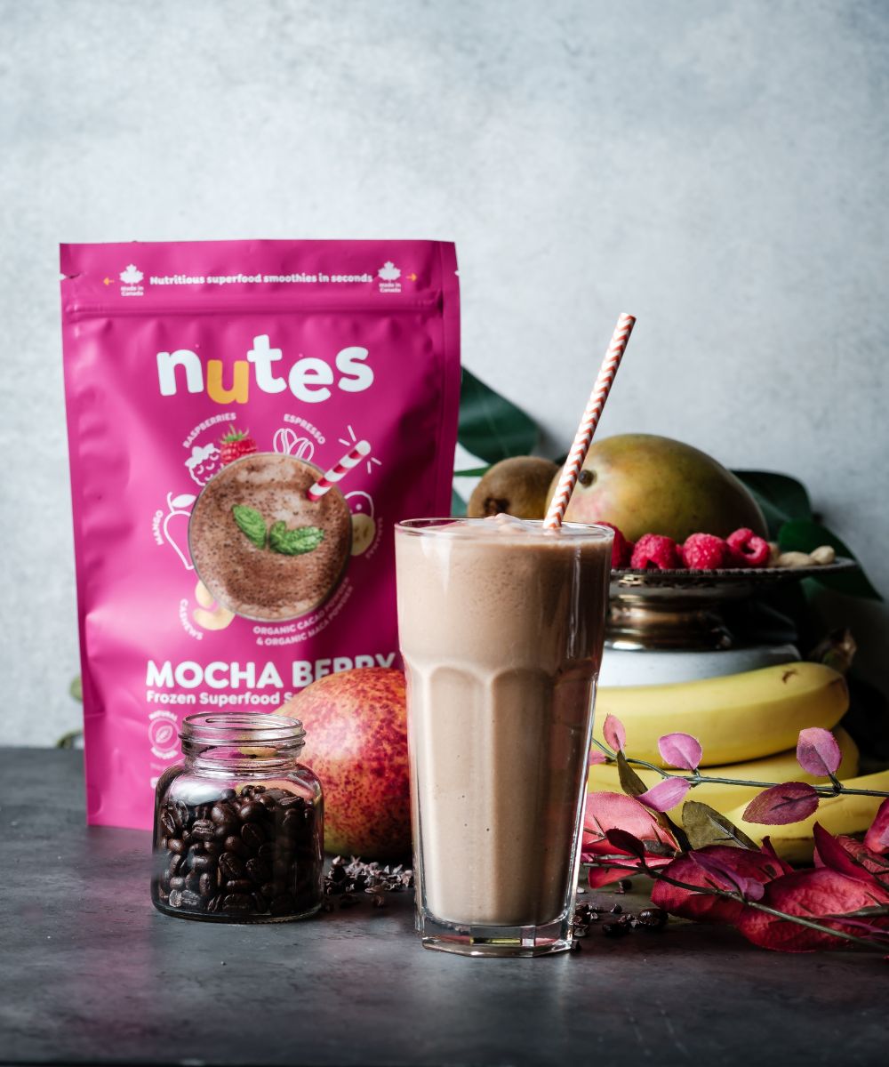 Nutes frozen superfood Mocha Berry Smoothie in glass with paper straw, surrounded by fresh fruit, beans, and product pouch.
