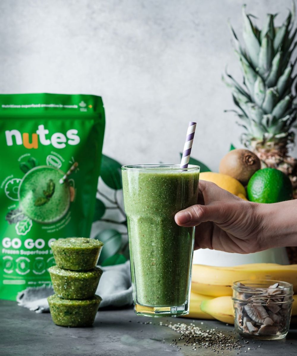 Nutes frozen superfood go go green Smoothie in glass with paper straw, surrounded by fresh fruit, smoothie pucks, and product pouch.